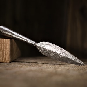 May include: A forged metal spearhead with a textured surface, resting on a wooden surface next to a rectangular wooden block. The spearhead is silver and has a sharp point. The background is dark.