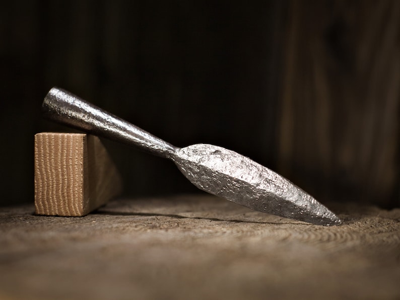 May include: A forged metal spearhead with a textured surface, resting on a wooden surface next to a rectangular wooden block. The spearhead is silver and has a sharp point. The background is dark.