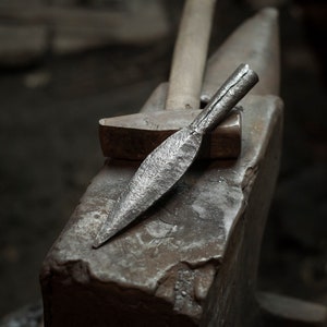 May include: A close-up of a metal spearhead resting on an anvil. The spearhead is made of steel and has a sharp point. The anvil is made of iron and has a flat surface.