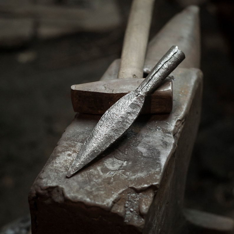 May include: A close-up of a metal spearhead resting on an anvil. The spearhead is made of steel and has a sharp point. The anvil is made of iron and has a flat surface.