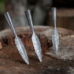 May include: Three handcrafted metal spearheads with a textured, silvery finish are displayed on a weathered wooden surface. The spearheads have a pointed, leaf-shaped blade and a cylindrical base.
