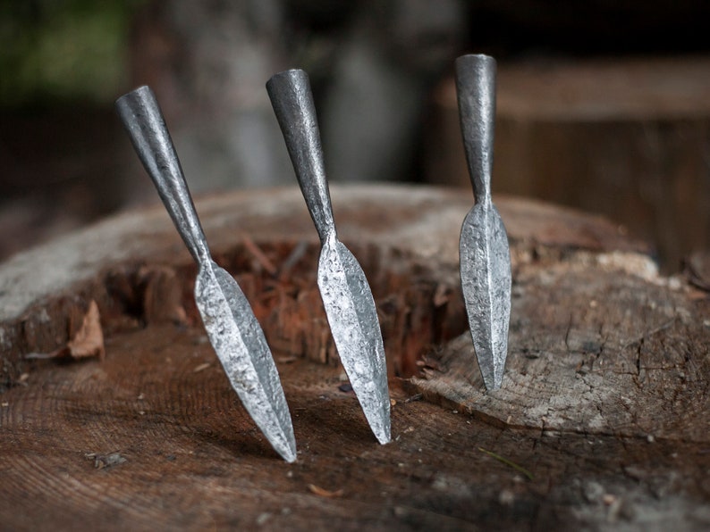 May include: Three handcrafted metal spearheads with a textured, silvery finish are displayed on a weathered wooden surface. The spearheads have a pointed, leaf-shaped blade and a cylindrical base.