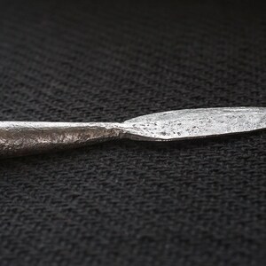May include: A forged metal spearhead with a textured surface. The spearhead has a cylindrical base that tapers to a sharp, pointed tip. The metal has a dark, aged appearance, suggesting it is an antique or historical artifact.