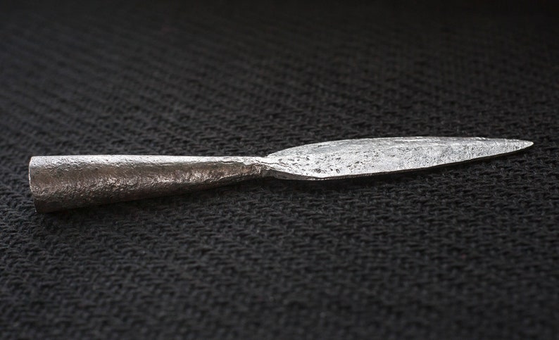 May include: A forged metal spearhead with a textured surface. The spearhead has a cylindrical base that tapers to a sharp, pointed tip. The metal has a dark, aged appearance, suggesting it is an antique or historical artifact.