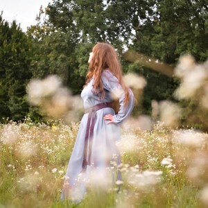 Op de afbeelding: Een vrouw met lang, rood haar draagt een lichtblauwe jurk met een donkerpaarse riem en staat in een veld met witte bloemen.