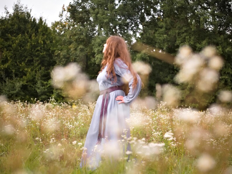 Op de afbeelding: Een vrouw met lang, rood haar draagt een lichtblauwe jurk met een donkerpaarse riem en staat in een veld met witte bloemen.