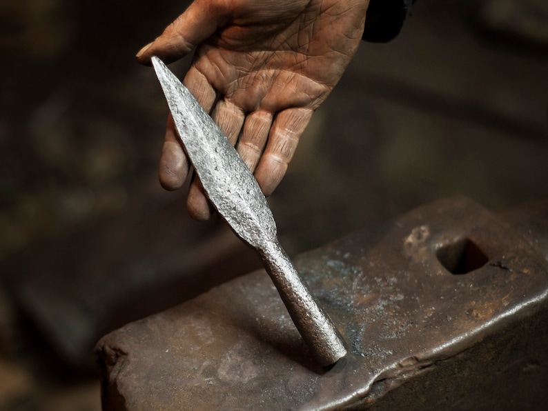 May include: A hand holds a silver spearhead, resting on a metal anvil. The spearhead is pointed and has a rough, textured surface.
