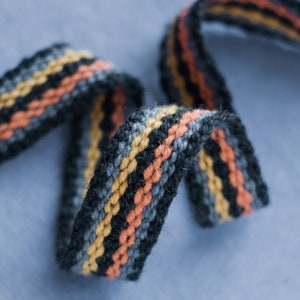 Peut inclure: Gros plan sur une sangle tissée noire, orange et jaune. La sangle est faite d'un matériau doux et texturé et présente un design tressé.
