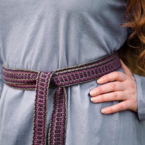 Op de afbeelding: Een langarmige, lichtblauwe tuniek met een geweven riem die om de taille is geknoopt. De riem is gemaakt van bruine, zwarte en paarse draden.