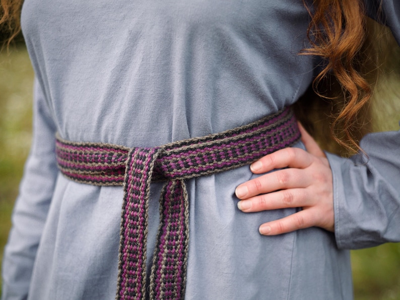 Op de afbeelding: Een langarmige, lichtblauwe tuniek met een geweven riem die om de taille is geknoopt. De riem is gemaakt van bruine, zwarte en paarse draden.