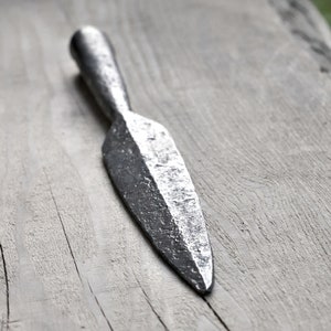 Może przedstawiać: Srebrna grot wł&oacute;czni z ostrym końcem i fakturowaną powierzchnią. Grot wł&oacute;czni leży na jasnoszarym drewnianym podłożu.
