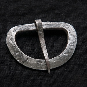 May include: A silver metal belt buckle with a simple, rounded design. The buckle has a single, straight pin.
