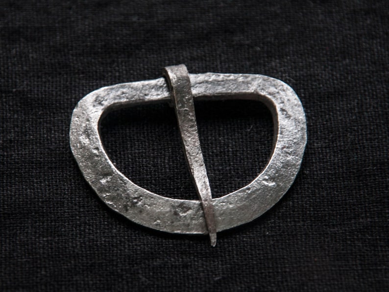May include: A silver metal belt buckle with a simple, rounded design. The buckle has a single, straight pin.
