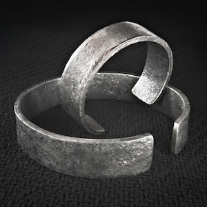 May include: Two silver-coloured metal cuff bracelets with a textured, hammered appearance. The bracelets are open-ended and rest on a dark, textured surface. The metal has a slightly aged, industrial look.