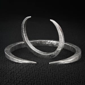 May include: Three silver-toned, open-ended bangles with a hammered texture. The bangles have a crescent shape, with the ends slightly curved. The background is a dark, textured surface, highlighting the jewelry's metallic sheen.