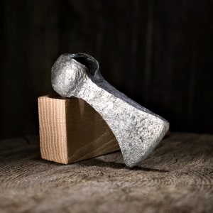 May include: A silver axe head with a rounded top and a slightly curved blade, resting on a wooden block.