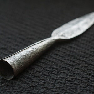 May include: A silver spearhead with a textured surface. The spearhead has a long, pointed blade and a cylindrical base. The metal has a slightly tarnished appearance, suggesting age or use. The spearhead is likely a historical artifact.
