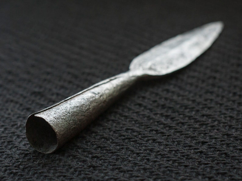 May include: A silver spearhead with a textured surface. The spearhead has a long, pointed blade and a cylindrical base. The metal has a slightly tarnished appearance, suggesting age or use. The spearhead is likely a historical artifact.
