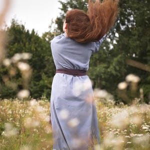 Op de afbeelding: Een vrouw met lang, rood haar staat in een veld met witte bloemen. Ze draagt een lichtblauwe jurk met een bruine riem. Haar haar waait in de wind.
