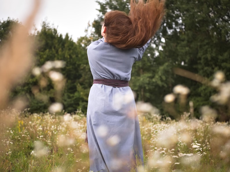 Op de afbeelding: Een vrouw met lang, rood haar staat in een veld met witte bloemen. Ze draagt een lichtblauwe jurk met een bruine riem. Haar haar waait in de wind.