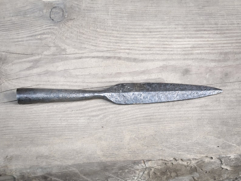 Może przedstawiać: Zbliżenie na metalową grot wł&oacute;czni z ostrym końcem i długim, wąskim ostrzem. Groty wł&oacute;czni są wykonane z ciemnego metalu i mają szorstką, fakturowaną powierzchnię.