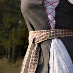 Puede incluir: Una túnica gris oscuro de estilo medieval con un cinturón tejido. La túnica presenta una parte delantera con cordones rojos y se usa sobre una prenda blanca. El cinturón tiene un patrón tejido marrón claro y gris.