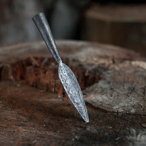May include: A hand-forged metal spearhead with a textured surface. The spearhead is silver-colored and has a leaf-like shape. It is resting on a weathered, brown wooden surface, suggesting a rustic or historical theme.