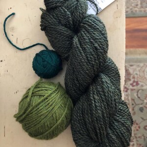 May include: An assortment of green yarn in various forms. A large skein of dark green yarn sits next to two smaller balls of yarn, one dark teal and one light green. A tag is visible in the upper right corner.