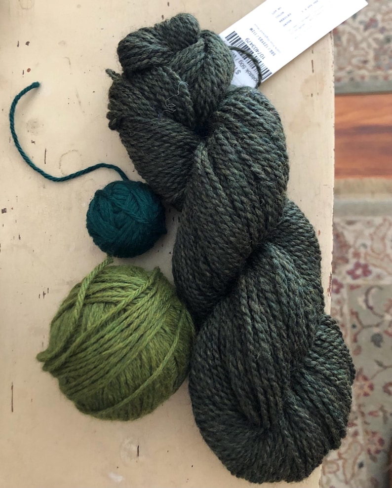 May include: An assortment of green yarn in various forms. A large skein of dark green yarn sits next to two smaller balls of yarn, one dark teal and one light green. A tag is visible in the upper right corner.