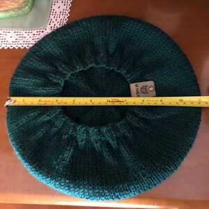 May include: A dark teal knitted beret, approximately 10 inches in diameter. The beret is made of a thick yarn and has a ribbed edge. A tag is attached to the beret. The beret is displayed on a wooden surface.