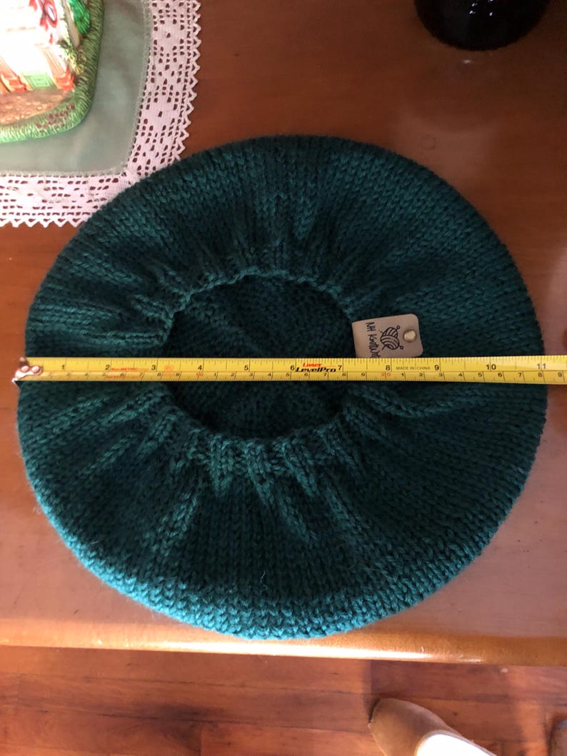 May include: A dark teal knitted beret, approximately 10 inches in diameter. The beret is made of a thick yarn and has a ribbed edge. A tag is attached to the beret. The beret is displayed on a wooden surface.