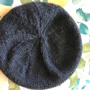 May include: A dark gray knitted beret. The beret is round with a ribbed texture and a central point where the knitting converges. The hat is made of a soft, possibly wool, material.