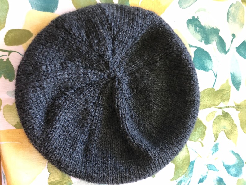 May include: A dark gray knitted beret. The beret is round with a ribbed texture and a central point where the knitting converges. The hat is made of a soft, possibly wool, material.