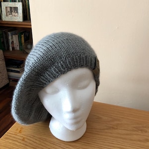 May include: A gray knit beret displayed on a white foam mannequin head. The beret has a ribbed texture and a small, brown leather tag. The hat is made of a soft, warm material, perfect for cold weather.