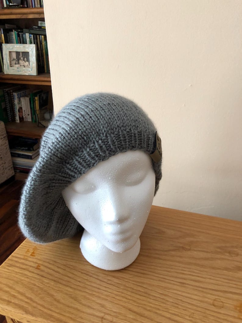 May include: A gray knit beret displayed on a white foam mannequin head. The beret has a ribbed texture and a small, brown leather tag. The hat is made of a soft, warm material, perfect for cold weather.