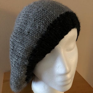 May include: A gray and black knit beret. The beret is displayed on a white mannequin head. The hat is made of a soft, textured yarn. The main body of the hat is gray, with a black band around the bottom.
