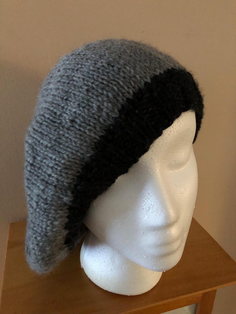 May include: A gray and black knit beret. The beret is displayed on a white mannequin head. The hat is made of a soft, textured yarn. The main body of the hat is gray, with a black band around the bottom.