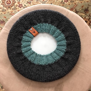 May include: A dark gray and teal knitted cowl with a circular design. The cowl features a teal ribbed section and a dark gray section. A small brown leather tag is attached to the cowl.