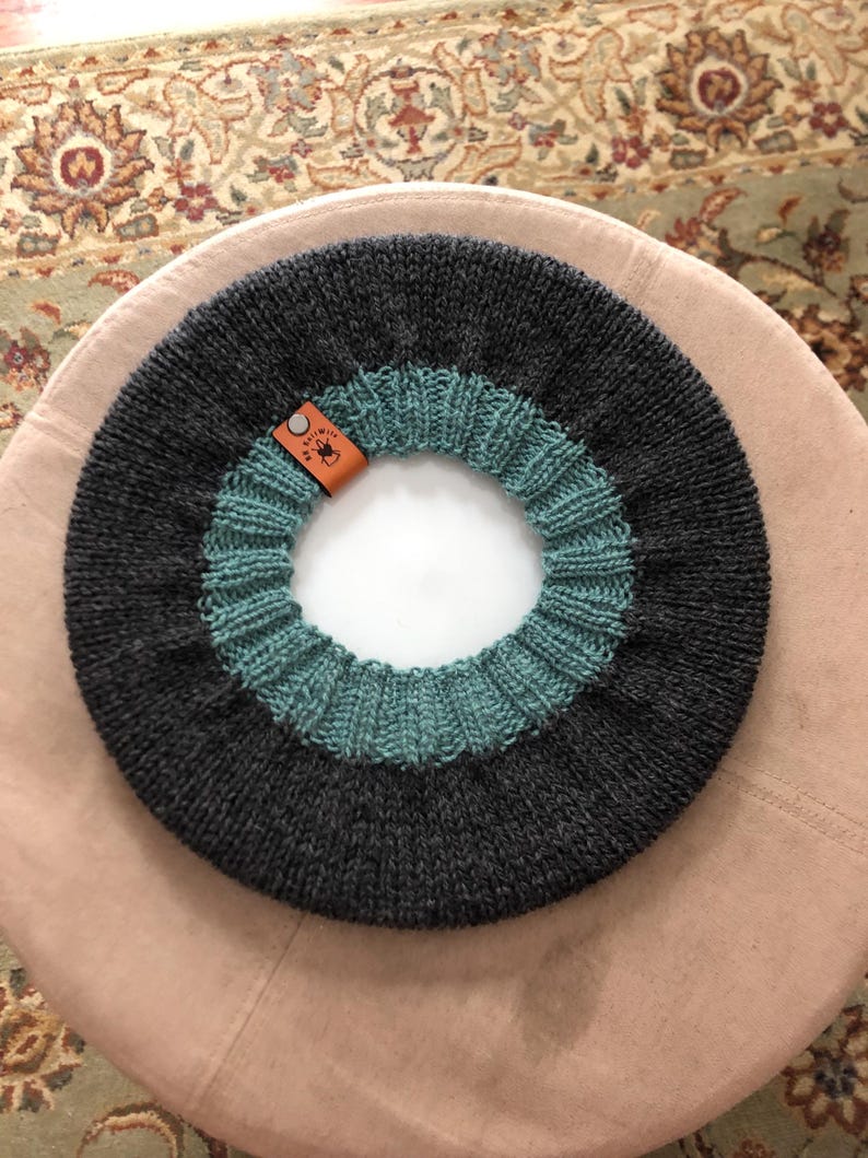May include: A dark gray and teal knitted cowl with a circular design. The cowl features a teal ribbed section and a dark gray section. A small brown leather tag is attached to the cowl.