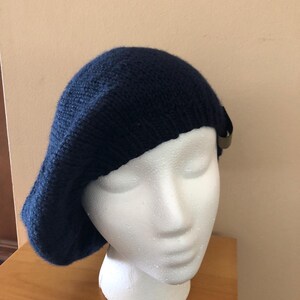 May include: A navy blue knit beret with a small, decorative accent. The beret is displayed on a white mannequin head, showcasing its shape and texture. The hat is made of a soft, textured material.