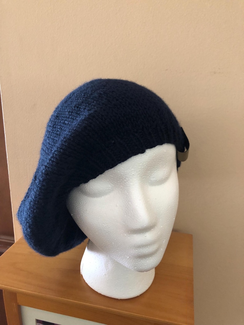 May include: A navy blue knit beret with a small, decorative accent. The beret is displayed on a white mannequin head, showcasing its shape and texture. The hat is made of a soft, textured material.
