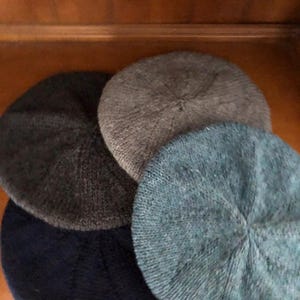 May include: Four knitted beret hats in shades of gray and blue are arranged in a close-up shot. The hats are circular with a ribbed texture, and the colors include dark gray, light gray, and two shades of blue.