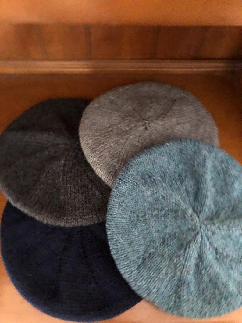 May include: Four knitted beret hats in shades of gray and blue are arranged in a close-up shot. The hats are circular with a ribbed texture, and the colors include dark gray, light gray, and two shades of blue.