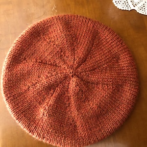 May include: A rust-colored knitted beret, circular in shape, with a textured, woven appearance. The beret is made of a soft, warm material, perfect for cooler weather. The hat is a solid color with a subtle pattern radiating from the center.
