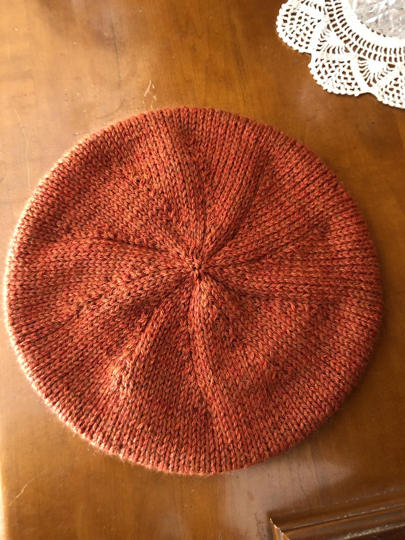 May include: A rust-colored knitted beret, circular in shape, with a textured, woven appearance. The beret is made of a soft, warm material, perfect for cooler weather. The hat is a solid color with a subtle pattern radiating from the center.