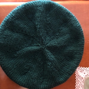 May include: A dark green knitted beret. The hat is round with a central point where the knitting converges. The beret is made of a textured yarn, and the color is a deep, forest green. The beret is a classic style, suitable for various occasions.