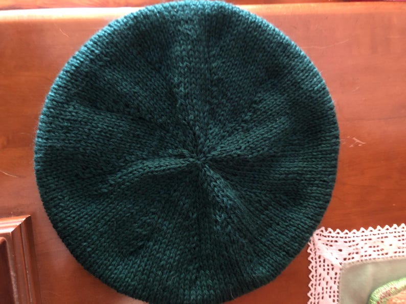 May include: A dark green knitted beret. The hat is round with a central point where the knitting converges. The beret is made of a textured yarn, and the color is a deep, forest green. The beret is a classic style, suitable for various occasions.