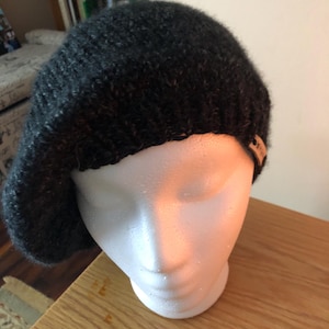 May include: A dark gray knitted beret displayed on a white mannequin head. The beret is made of a textured yarn and has a small tag attached. The beret is a classic style, suitable for various occasions.