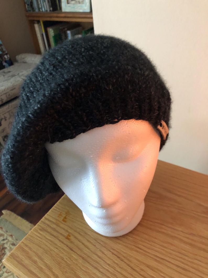 May include: A dark gray knitted beret displayed on a white mannequin head. The beret is made of a textured yarn and has a small tag attached. The beret is a classic style, suitable for various occasions.