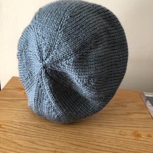 May include: A knitted, light blue beret hat. The hat is made of a soft, textured yarn and has a rounded crown. The beret is resting on a wooden surface.